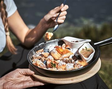A narrow gravel path winds through an autumn forest. Colorful leaves and tall trees surround the landscape, while mountains can be seen in the background.