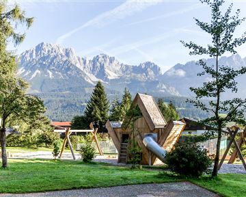 A beautiful playground in a park with a unique play equipment and shade-giving trees. In the background, impressive mountains are visible.