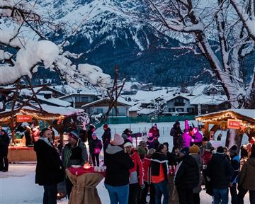 Ein winterlicher Weihnachtsmarkt mit Menschen in warmen Kleidern. Im Hintergrund sind schneebedeckte Berge zu sehen.