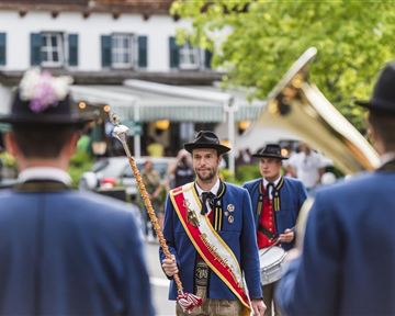 A traditional costume parade with people in blue jackets and hats. In the foreground stands a man with a scepter and an honorary ribbon.