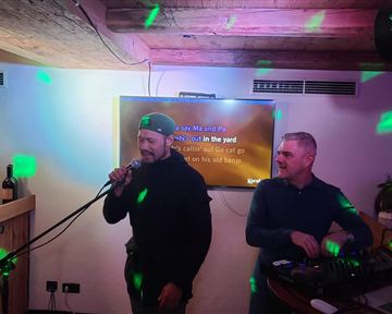 Zwei Männer stehen in einer Bar. Einer singt mit einem Mikrofon, während der andere am Mischpult arbeitet.