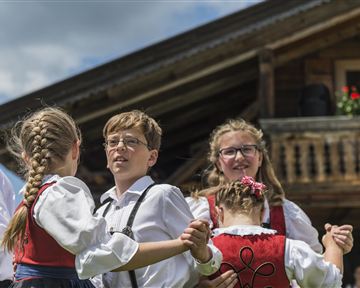 Children in traditional costumes are dancing together in front of a rustic wooden house. The sky is blue and the atmosphere is cheerful.