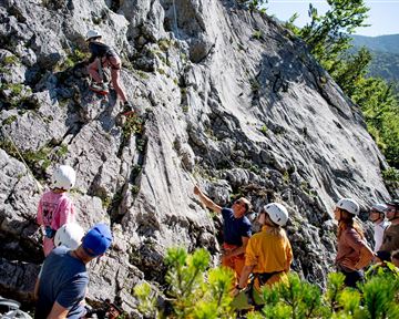 Eine Gruppe von Menschen beobachtet einen Kletterer an einer Felswand. Der Hintergrund zeigt grüne Bäume und Berge im Freien.
