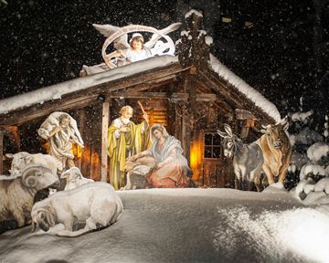 Eine winterliche Krippe mit Maria, Josef und dem neugeborenen Jesus in einem Stall, umgeben von Tieren. Sanfte Schneeflocken fallen auf die Szene und erzeugen eine festliche Atmosphäre.