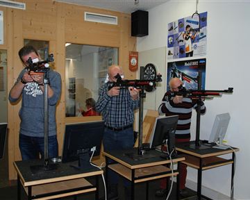 Drei Männer stehen an Schießständen mit Gewehren. Sie konzentrieren sich auf das Zielen und Schießen, während Computer vor ihnen stehen.