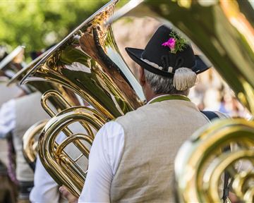 A group of musicians with tubas is wearing traditional clothing. In the background, trees and a festive atmosphere can be seen.