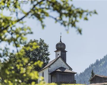 Eine malerische Kirche umgeben von Bäumen und Bergen. Der Himmel ist klar und blau.