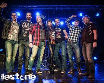 A group of seven musicians is standing on a stage. They are smiling and posing in front of a festive background with blue lights.