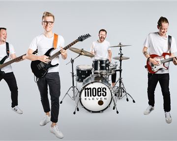 Eine Band mit vier Mitgliedern steht in einem hellen Raum. Sie spielen verschiedene Instrumente und lächeln in die Kamera.