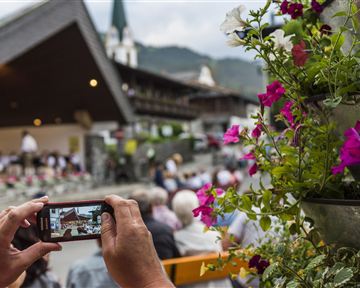 A person is holding a cellphone and photographing an outdoor event. In the foreground, colorful flowers are visible, while in the background, the crowd and a mountainous landscape can be seen.