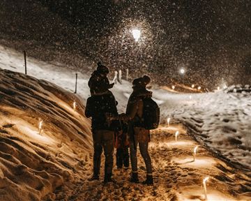 A family strolls through the snow at night as snowflakes fall. The path is lined with lights that create a festive atmosphere.