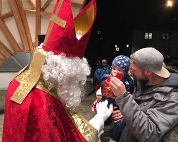 Ein verkleideter Weihnachtsmann spricht mit einem Vater und seinem kleinen Kind. Im Hintergrund sind weitere Personen und eine festliche Atmosphäre zu sehen.