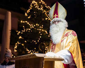 A man in religious garb with a white beard stands in front of a Christmas tree. It is a festive atmosphere with lights in the background.