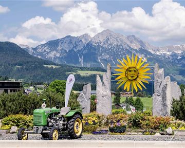 A green tractor stands in front of a sunny, blooming flower bed. In the background, majestic mountains and a bright blue sky can be seen.