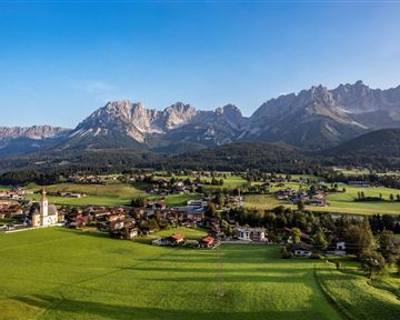 A picturesque landscape with green meadows and a small village. In the background, majestic mountains rise under a clear sky.