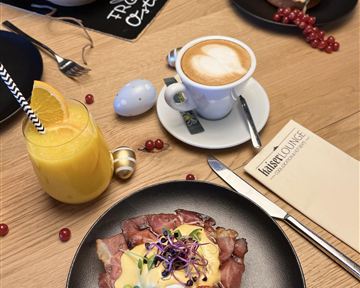 Ein köstliches Frühstück mit Rinderbraten, pochiertem Ei und Kirschtomaten auf einem schwarzen Teller. Im Hintergrund sind eine Tasse Kaffee und ein Glas Orangensaft zu sehen.