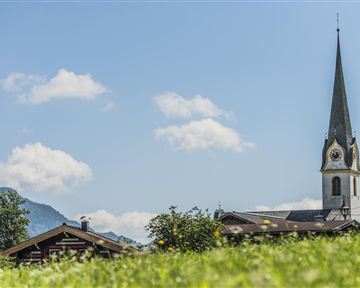 A picturesque landscape with a church and a traditional building in the foreground. The sky is blue with some clouds and the mountains are visible in the background.