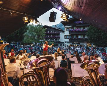 Ein Open-Air-Konzert mit einem Orchester auf einer Bühne. Viele Zuschauer genießen die Aufführung im Freien.