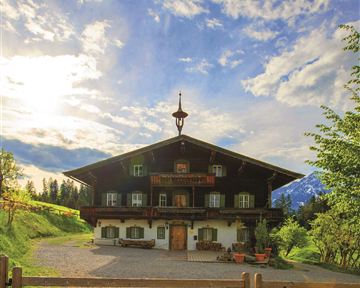 A traditional alpine house with light wood and a beautiful garden. The surroundings are green and surrounded by mountains.