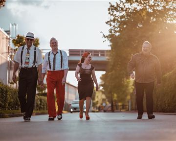 Four people are walking together on a street, surrounded by trees and buildings. The sun is shining, creating a warm atmosphere.