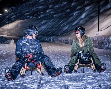 Zwei Personen sitzen auf Schlitten im Schnee bei Nacht. Die Umgebung ist beleuchtet und es ist eine winterliche Atmosphäre sichtbar.