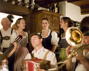 Eine fröhliche Gruppe von Menschen in traditioneller Tracht singt und spielt Musik in einem rustikalen Raum. Es gibt Instrumente wie Gitarre und Trompete, die zur festlichen Stimmung beitragen.