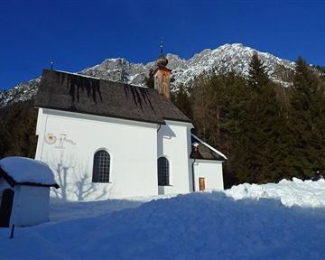 Eine kleine weiße Kapelle steht im Schnee, umgeben von hohen Bergen und Nadelbäumen. Der klare blaue Himmel bietet einen schönen Kontrast zur winterlichen Landschaft.