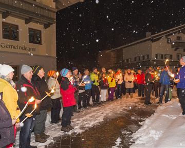 A group of people is standing in the snow and holding torches. It is snowing, and the scene takes place at night.