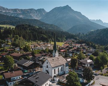 A picturesque village view with a white church in the foreground and the Alps in the background. The surroundings are green and mountainous, ideal for nature lovers.