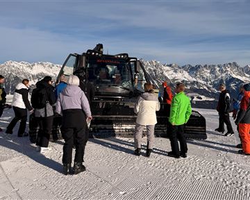 A group of people is standing around a snow groomer in the mountains. In the background, snow-covered peaks and a clear sky can be seen.