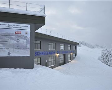 Ein modernes Gebäude in einer verschneiten Landschaft. Schilder mit technischen Informationen zur Schneeproduktion sind sichtbar.