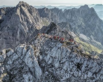 A breathtaking view of a rugged mountain ridge with many people on the summit. The landscape is surrounded by majestic mountains and a clear expanse of sky.