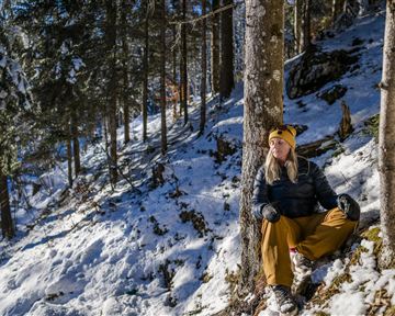 Eine Person sitzt im Schnee, gestützt an einem Baum. Umgeben von verschneiten Bäumen und einer klaren Winterlandschaft.