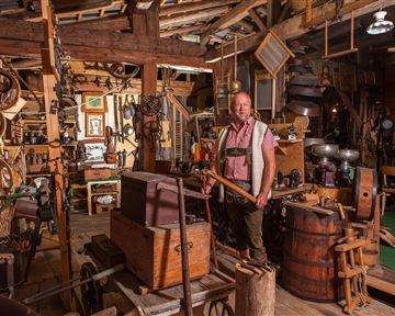 A man stands in an old craft room full of tools and memorabilia. The atmosphere is rustic and traditional.