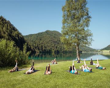 Eine Gruppe von Menschen macht Dehnübungen auf einer grünen Wiese neben einem klaren See. Im Hintergrund sind bewaldete Hügel und ein blauer Himmel zu sehen.