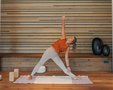 Eine Frau macht Yoga in einem hellen Raum mit Holzverkleidung. Sie befindet sich in der Position des Kriegers und wirkt konzentriert und entspannt.