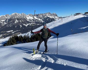 Ein Skifahrer steht im Schnee und posiert mit einem Skistock. Im Hintergrund sind schneebedeckte Berge und ein blauer Himmel zu sehen.