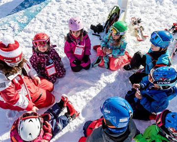 Eine Gruppe von Kindern sitzt im Schnee und hört einem Skilehrer zu. Rundherum sind Skiausrüstungen und verschneite Berge sichtbar.