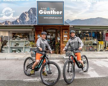Zwei Radfahrer stehen mit ihren Fahrrädern vor einem Sportgeschäft. Im Hintergrund sind die Berge und das Geschäftsschild sichtbar.