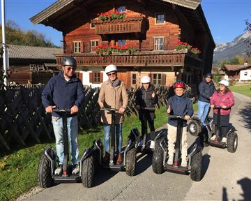 Eine Gruppe von sechs Personen fährt auf Segways in ländlicher Umgebung. Im Hintergrund ist ein traditionelles Holzhaupthaus mit Blumen zu sehen.