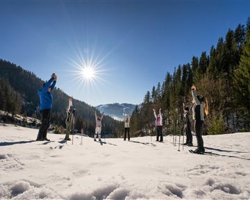 Eine Gruppe von Menschen macht eine Dehnübung im Schnee. Die Sonne scheint hell in den klaren blauen Himmel.