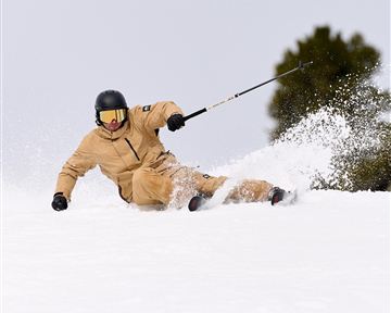 Ein Skifahrer fährt in einem verschneiten Gebiet. Er trägt eine beige Skijacke und einen Helm.