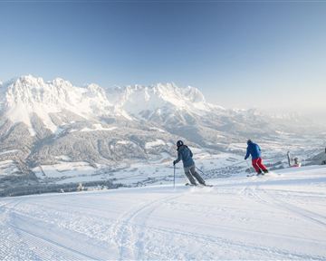 Two skiers are skiing down the snowy slope. In the background, majestic mountains and a clear sky can be seen.