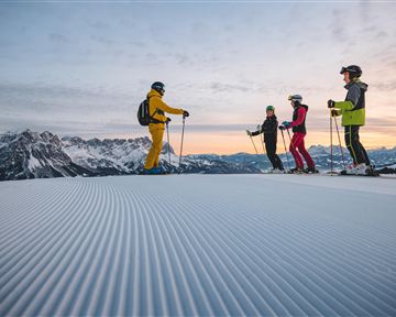 Eine Gruppe von Skifahrern steht auf einer frisch präparierten Piste in den Bergen. Die Aussicht zeigt schneebedeckte Gipfel und einen klaren Himmel bei Sonnenaufgang.