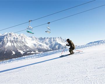 Ein Skifahrer fährt auf einer verschneiten Piste mit beeindruckenden Bergen im Hintergrund. Im Himmel sind Skilifte zu sehen.