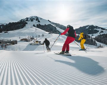 Drei Skifahrer fahren die verschneite Piste hinunter. Im Hintergrund sind schneebedeckte Berge und eine klare Winterlandschaft zu sehen.
