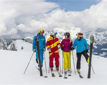 Eine Gruppe von vier Skifahrern steht auf einer verschneiten Piste. Im Hintergrund sind Berge und ein bewölkter Himmel zu sehen.
