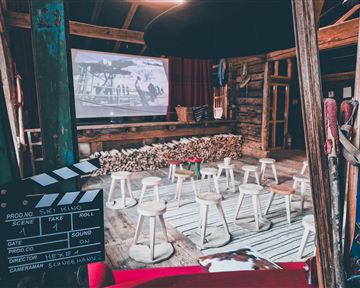 Ein gemütlicher Veranstaltungsraum mit Stühlen und einem großen Bildschirm. Der Raum hat eine rustikale Atmosphäre mit Holzelementen und einer Filmklappe im Vordergrund.