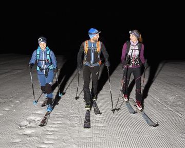 Drei Skifahrer gehen nachts über einen schneebedeckten Hang. Sie tragen Stirnlampen und skifahren in einem tiefen, dunklen Umfeld.
