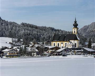 Eine schneebedeckte Landschaft mit einem charmanten Dorf und einer Kirche im Vordergrund. Im Hintergrund sind verschneite Berge und ein klarer Himmel zu sehen.
